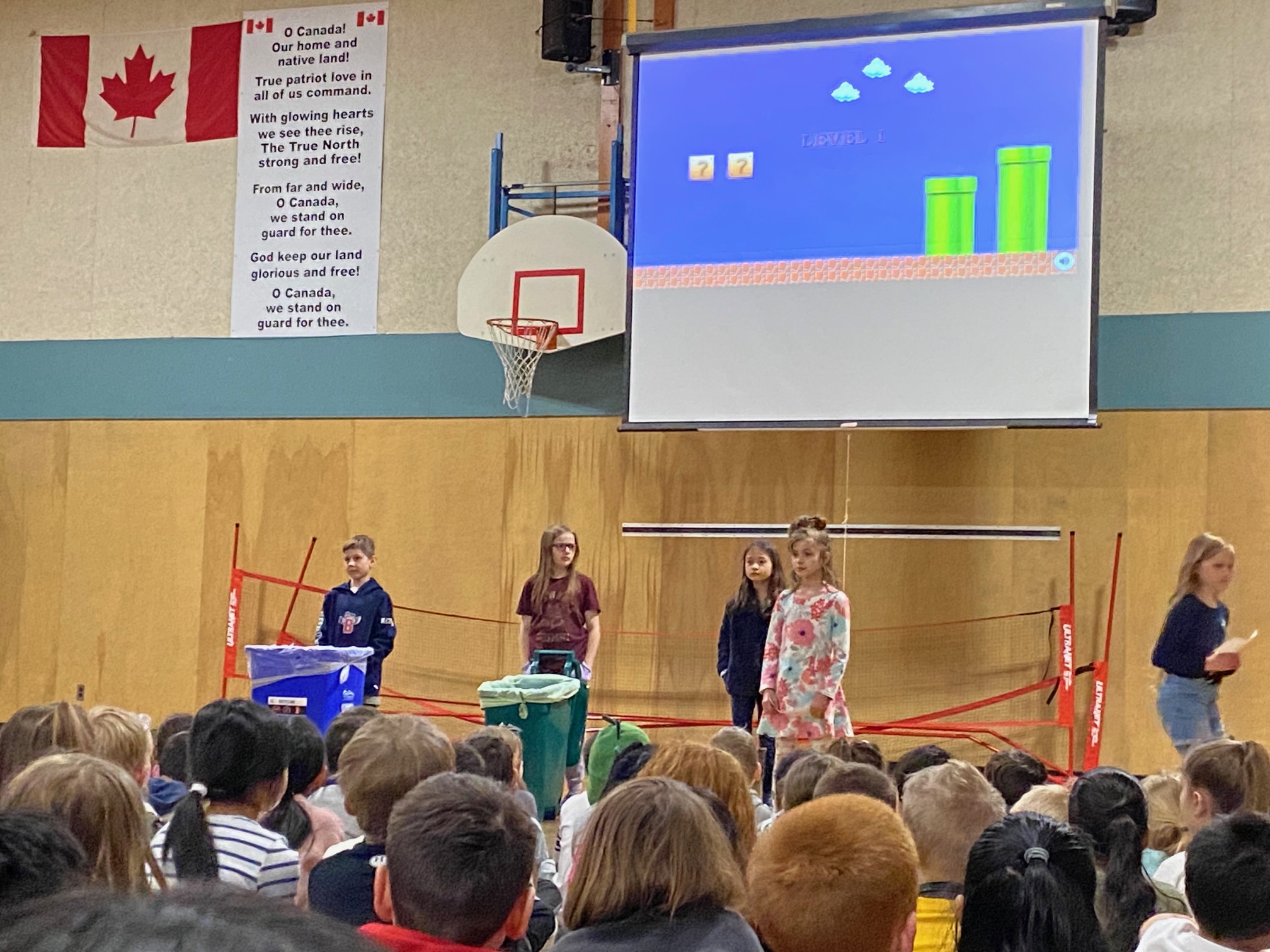 Students from a Grade 3/4 class at Highland Park Elementary stand behind garbage, organics and recycling bins at the front of the gymnasium during a school-wide assembly.