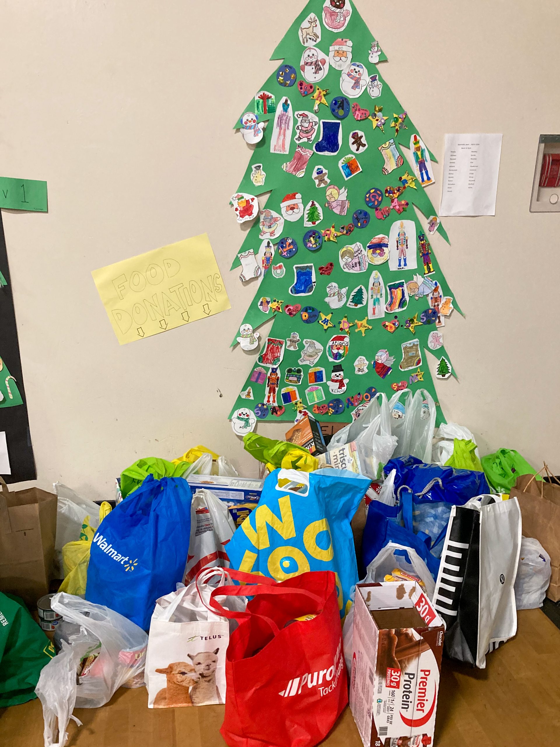 Food drive items in shopping bags underneath a Christmas tree made of poster at Eric Langton Elementary.