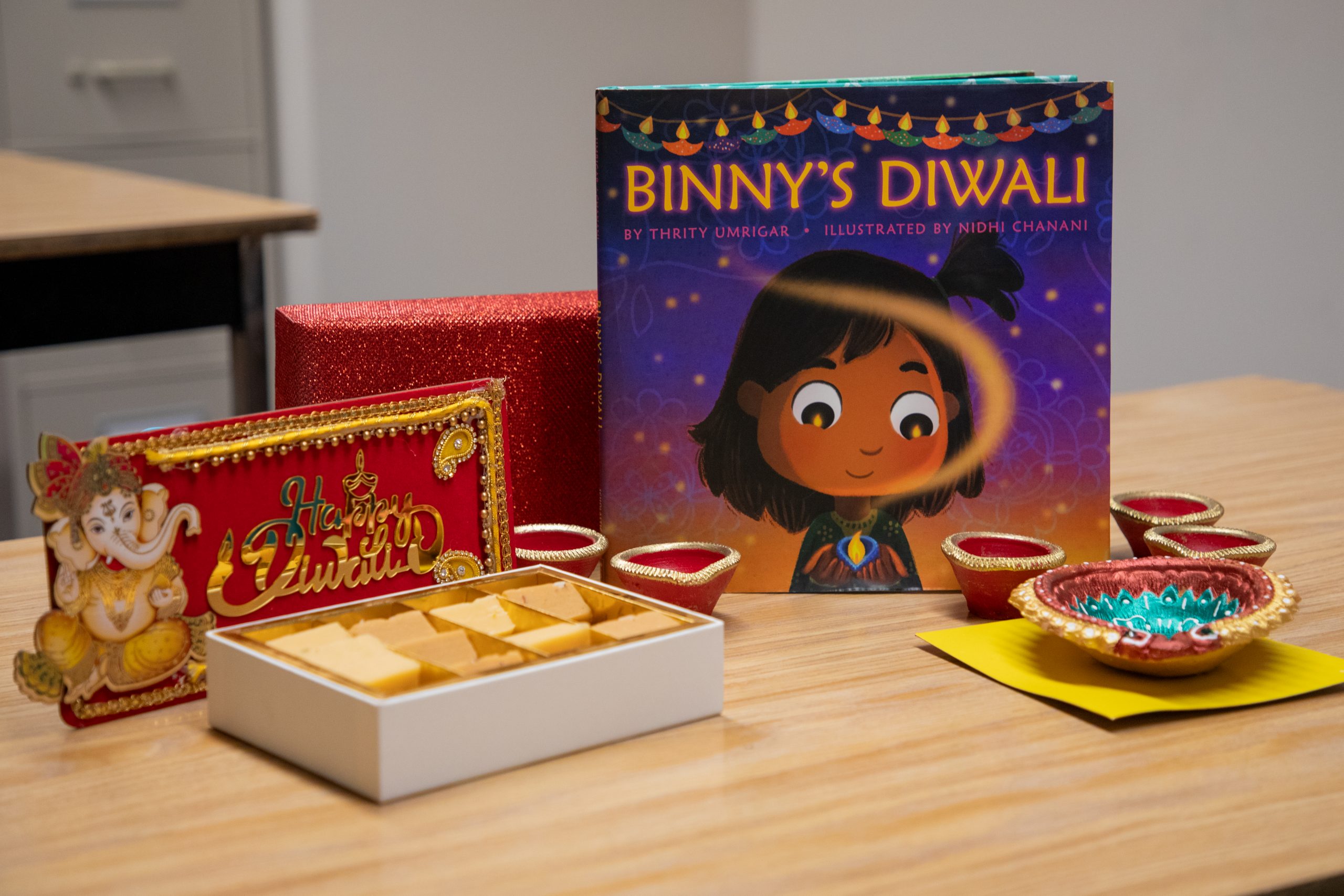 The book, "Binny's Diwali" by Thrity Umrigar, on a table surrounded by diyas, sweets and a sign that reads "Happy Diwali." 